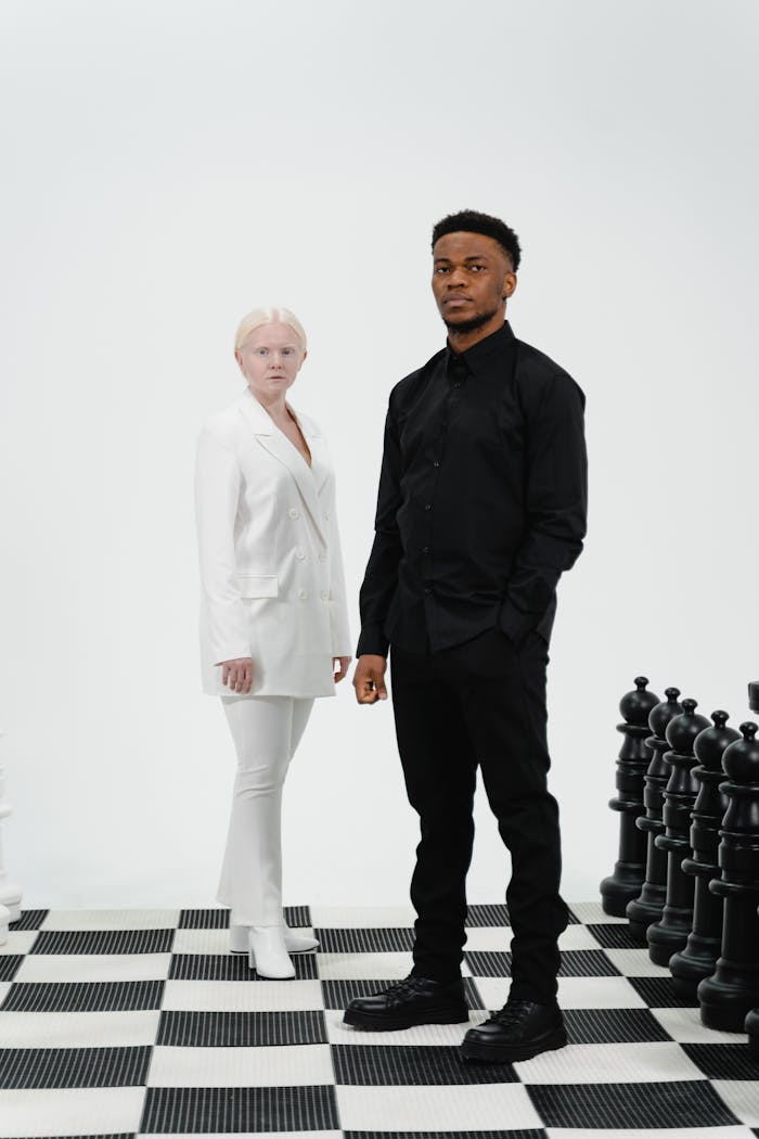 Two individuals in contrasting outfits stand on a giant chessboard with pieces.